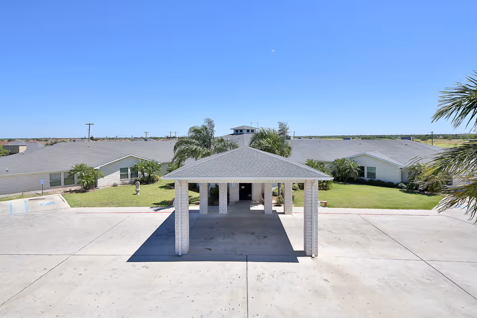 Front entrance canopy and driveway of a low-rise nursing facility with manicured lawns and palm trees under a clear blue sky.