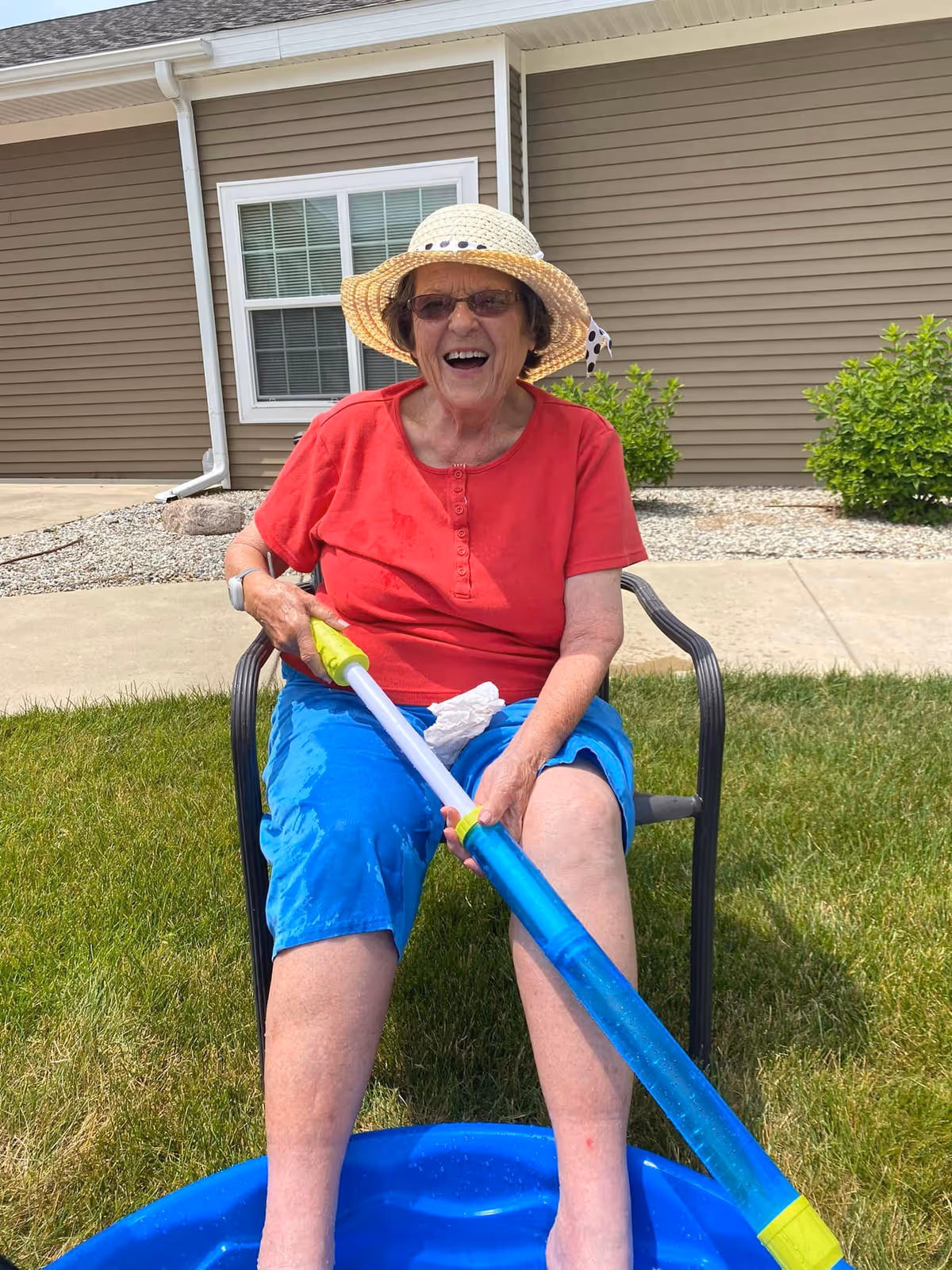 An elderly woman wearing a straw hat, sunglasses, a red shirt, and blue shorts is sitting on a black metal chair on green grass outside a building with beige siding. She is smiling and holding a blue and yellow water toy, with her feet in a small blue plastic pool.