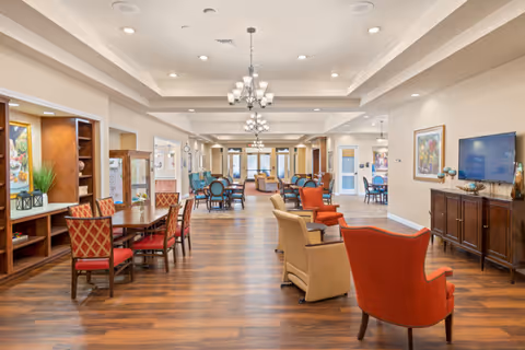 A spacious and well-lit senior living common area with wooden flooring, multiple seating arrangements including red and beige armchairs, and dining tables with red cushioned chairs. The room features chandeliers, built-in wooden shelves with decorative items, framed artwork on the walls, and a flat-screen TV on a wooden cabinet.