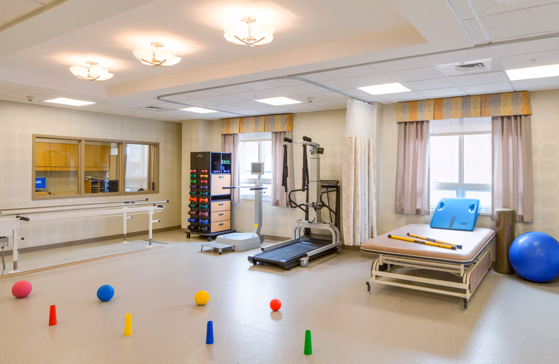 A bright physical therapy room with exercise equipment including a treadmill, balance platform, parallel bars, dumbbells, therapy balls, and a treatment table. The room has large windows with curtains and soft overhead lighting.