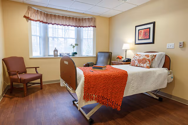 A cozy bedroom in a senior living facility with a single bed covered in a white blanket and an orange throw. There is a decorative pillow and a framed floral picture above the bed. Next to the bed is a nightstand with a lamp and a small flower arrangement. Two chairs are positioned near a window with a valance curtain, and the window sill has decorative items including a 'WELCOME' sign.