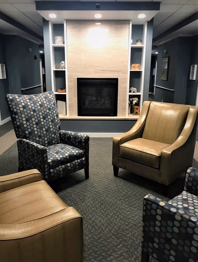 A cozy seating area with two patterned armchairs and two tan leather armchairs arranged around a modern fireplace with built-in shelves on either side, in a softly lit hallway with dark walls and carpeted floor.
