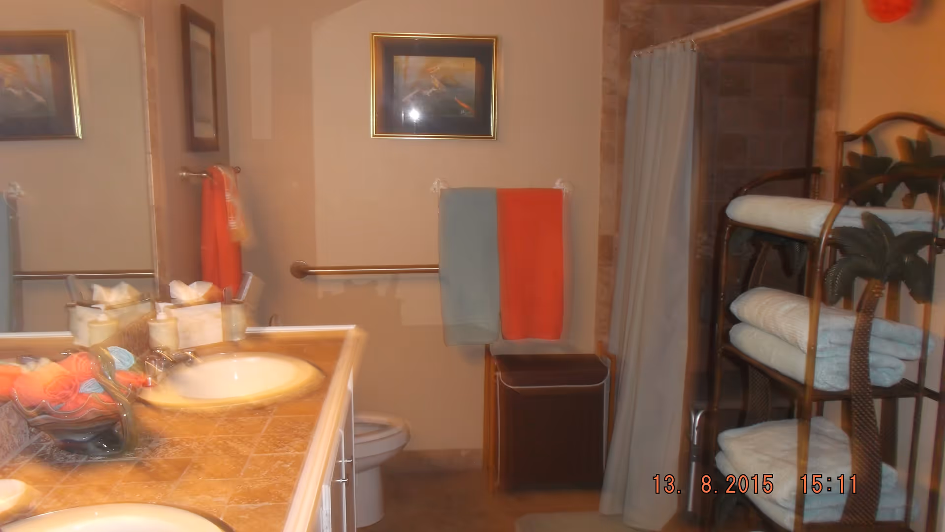 Bathroom with a double sink vanity topped with a tiled countertop, a large mirror above the sinks, a toilet, a towel rack with blue and orange towels, a shower with a curtain, and a shelving unit holding neatly folded white towels.