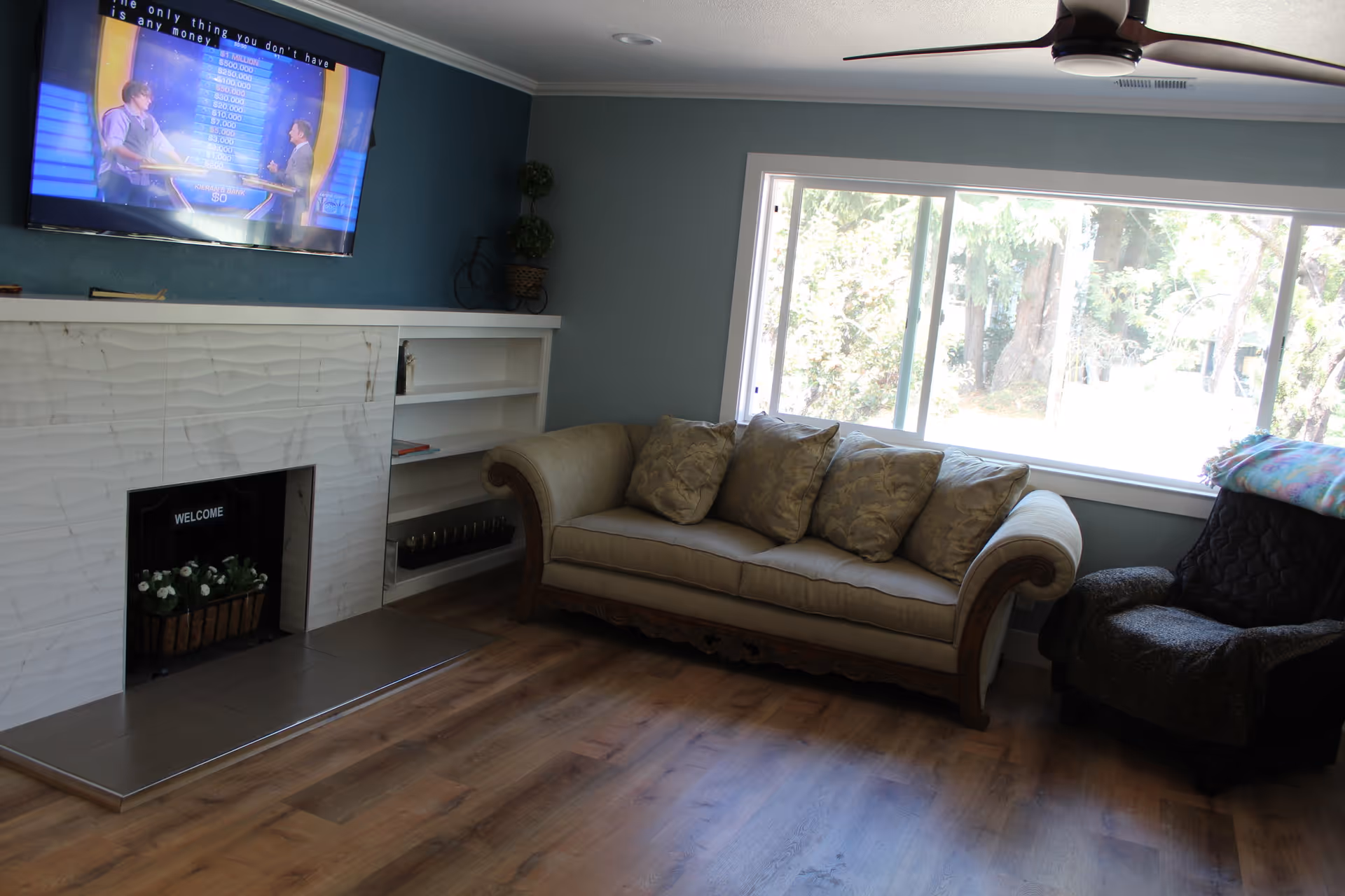 A cozy living room with a beige sofa adorned with patterned cushions, a dark armchair with a colorful blanket draped over the back, a large window letting in natural light, a mounted flat-screen TV displaying a game show, and a white marble fireplace with a small plant inside and built-in shelves beside it.