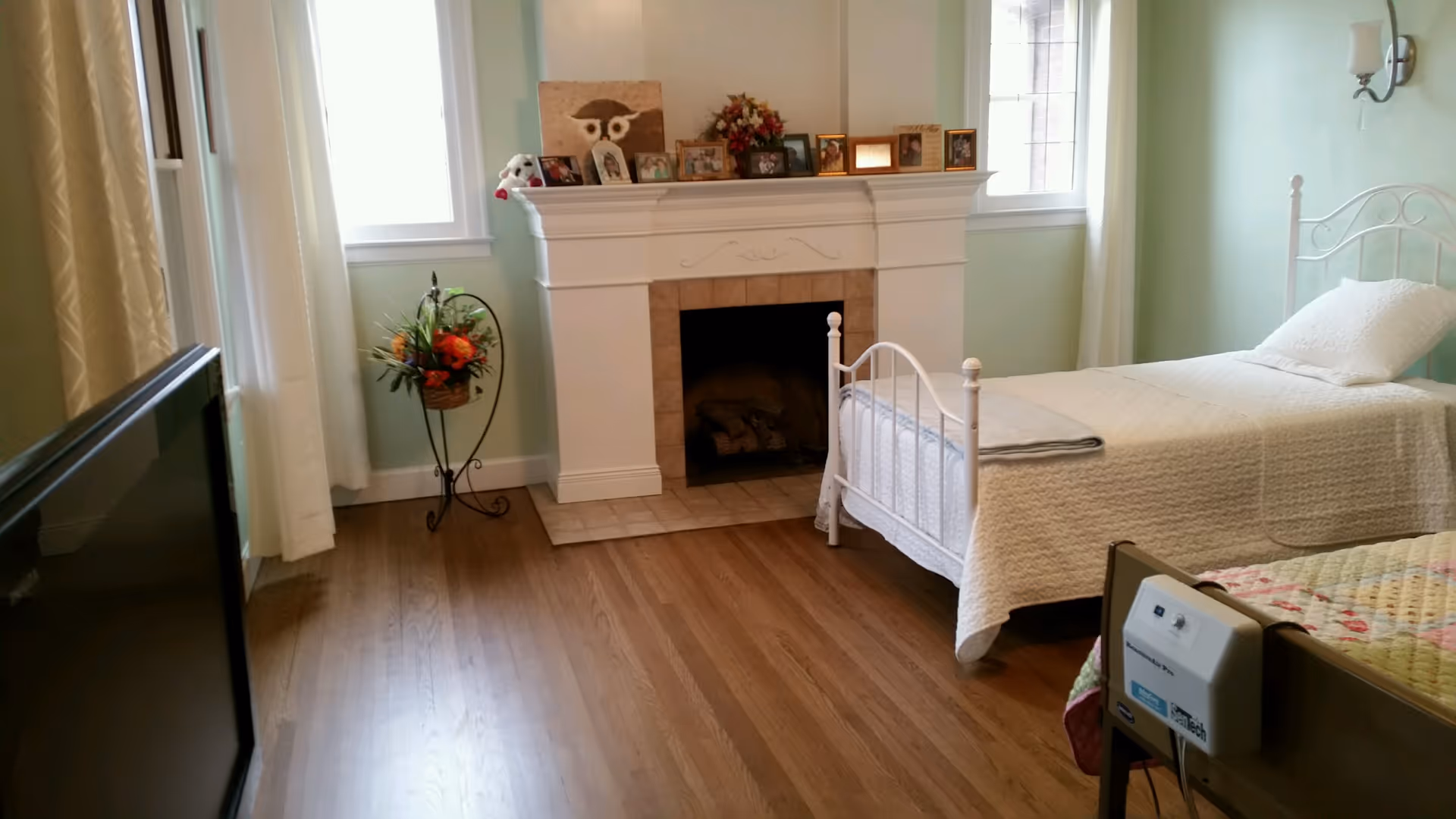 A cozy bedroom in a senior living facility featuring two beds with white metal frames and white bedding. There is a fireplace with a mantel decorated with framed photos and a flower arrangement. The room has light green walls, two windows with white curtains, a wooden floor, and a TV on the left side.