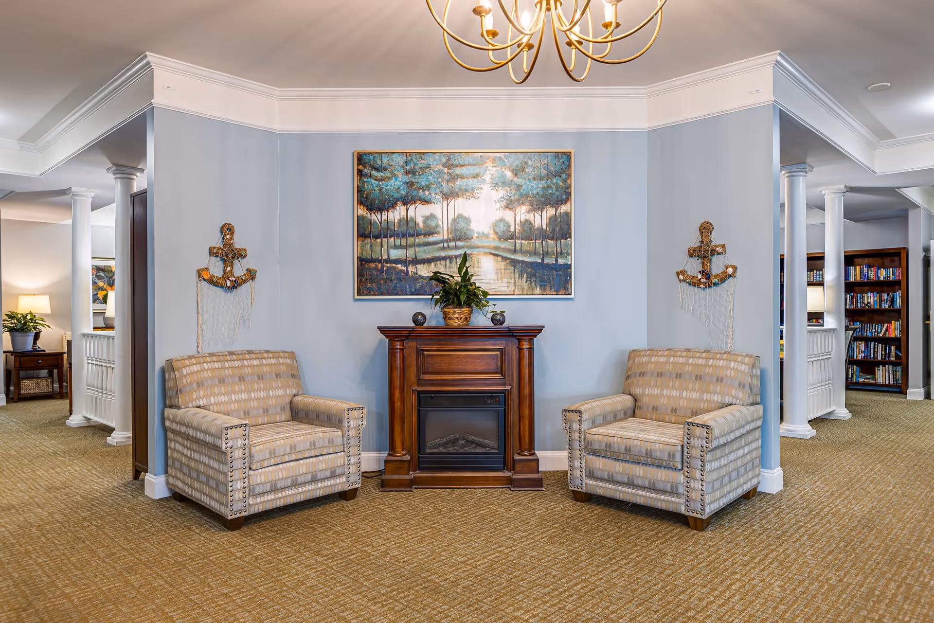 A cozy sitting area with two patterned armchairs facing each other, separated by a wooden electric fireplace. Above the fireplace hangs a large painting of a serene landscape with trees and a river. The walls are painted light blue, and there are decorative anchors with nets on either side of the fireplace. The carpet is beige, and white columns and bookshelves are visible in the background.
