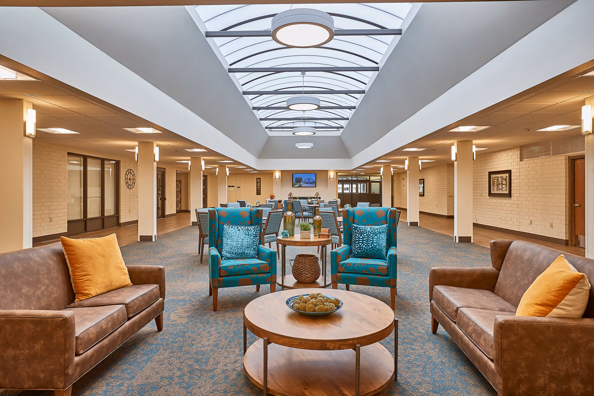 A spacious, well-lit common area with a high arched skylight ceiling. The room features two brown leather sofas with yellow and beige pillows, two blue patterned armchairs with matching pillows, and a round wooden coffee table with decorative items. In the background, there are several dining tables and chairs, wall sconces, framed artwork, and a TV mounted on the far wall.