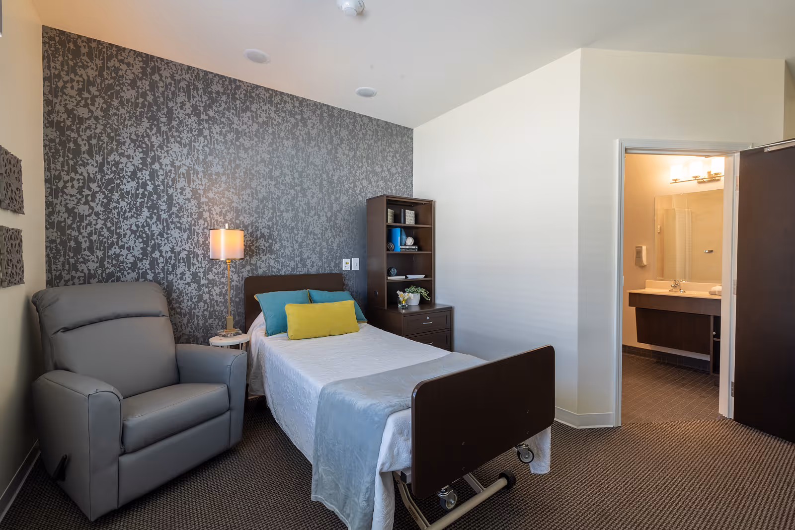 A senior living facility bedroom with a single bed covered in white and light blue bedding, accented with blue and yellow pillows. Next to the bed is a small round table with a lamp and a gray recliner chair. Behind the bed is a dark patterned accent wall. To the right, there is a dark wooden bookshelf and dresser. An open door reveals a bathroom with a sink, mirror, and lighting fixtures.