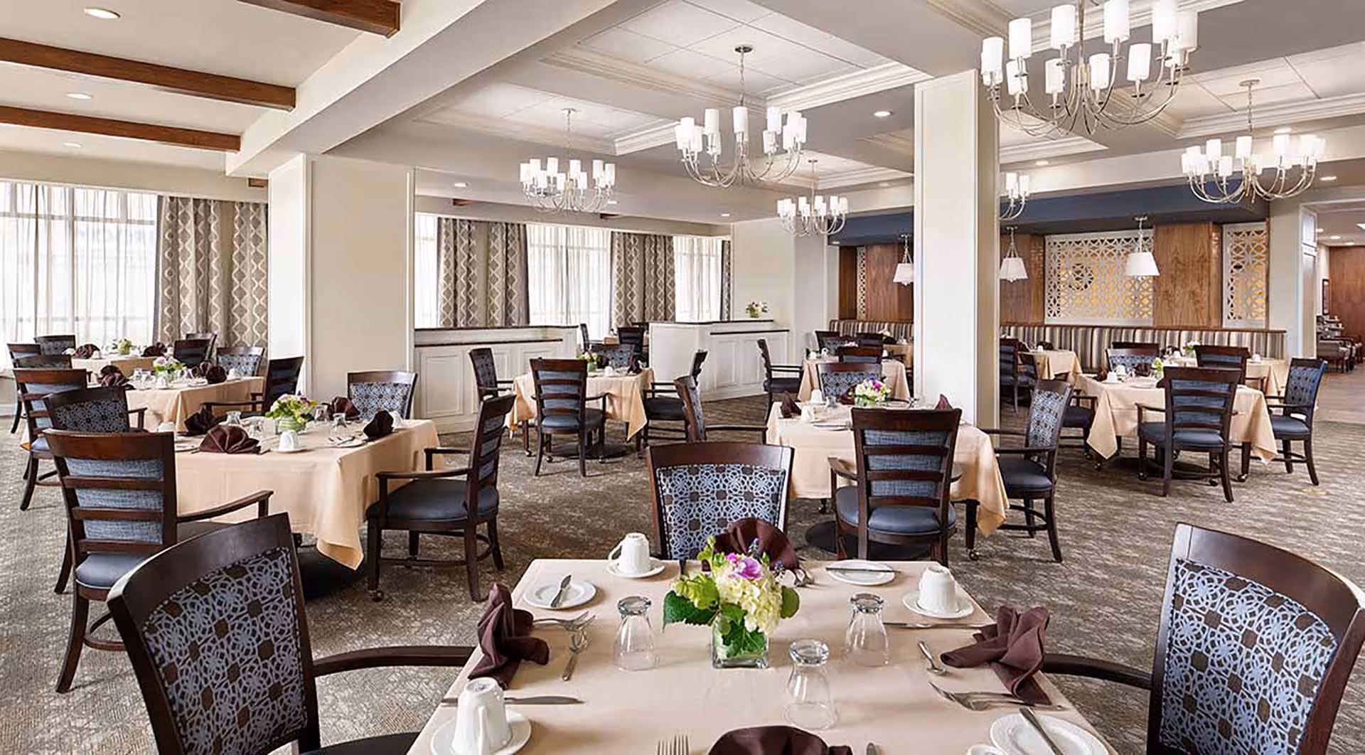 A spacious and elegant dining room with multiple tables covered in beige tablecloths, each set with white cups, plates, silverware, and brown folded napkins. The chairs have dark wooden frames with patterned upholstery. The room features large windows with patterned curtains, chandeliers hanging from the ceiling, and decorative wooden panels on the walls.