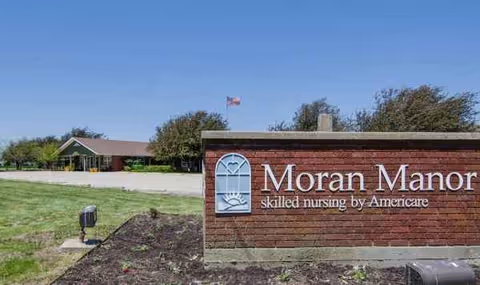Brick Moran Manor sign in foreground with facility building and clear blue sky.