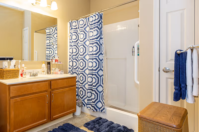 Bathroom with a wooden vanity cabinet, a large mirror, a sink with various toiletries on the counter, a shower with a blue and white patterned curtain, blue bath mats on the floor, and towels hanging on a rack attached to a white door.