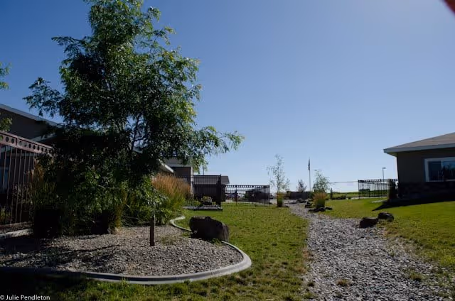 Outdoor garden area at DeSano Place Village with a gravel pathway, green grass, trees, and shrubs under a clear blue sky. Buildings are visible on either side of the pathway.