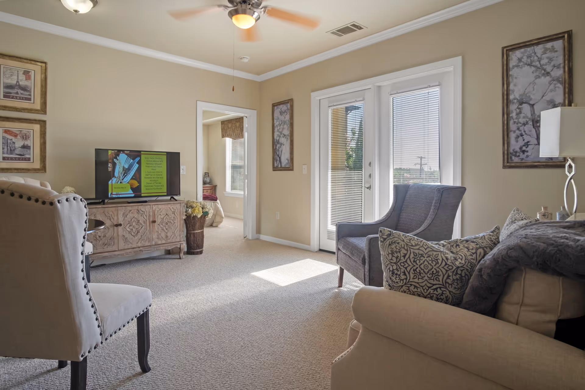 Sunlit living room with upholstered chairs, a decorative TV console, and glass French doors with blinds.