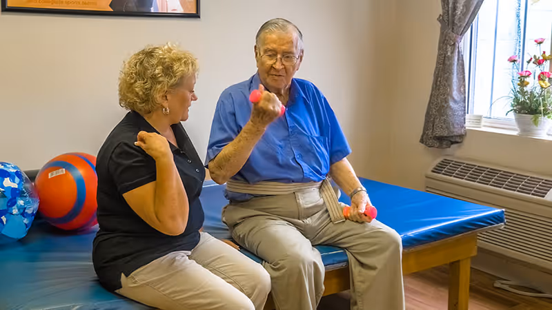 An elderly man in a blue shirt and beige pants is sitting on a padded exercise table holding small pink dumbbells, while a woman in a black shirt and beige pants sits beside him, assisting with the exercise. The room has a window with curtains and a flower pot on the windowsill.