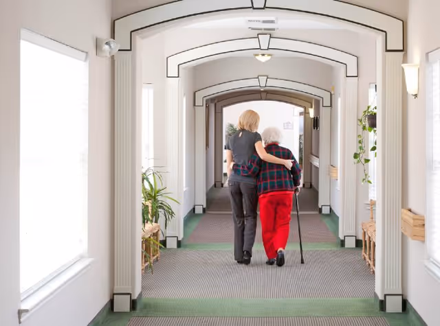 A caregiver walking arm-in-arm with an elderly woman using a cane down a well-lit hallway with arched ceilings and plants along the sides.