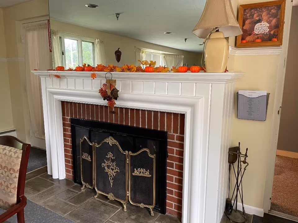 A cozy indoor fireplace with a white wooden mantel decorated with small orange pumpkins and autumn leaves. There is a decorative black and gold fireplace screen in front of the brick fireplace. To the right of the fireplace is a beige lamp on the mantel and a framed picture of pumpkins on the wall. A metal fireplace tool set is placed on the floor next to the fireplace. A chair with patterned upholstery is partially visible on the left side.