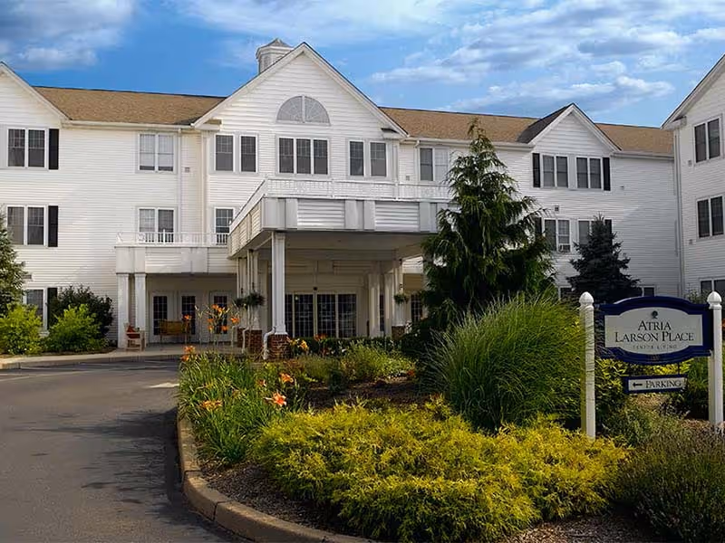 Exterior view of a large white senior living facility building with multiple windows and a covered entrance. There is a landscaped garden with green shrubs and orange flowers in front, and a sign that reads 'Atria Larson Place' with an arrow pointing to parking. The sky is clear and blue.