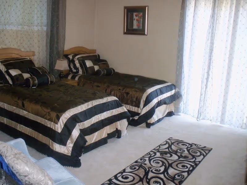 A bedroom with two single beds side by side, each with matching black and beige striped bedding and decorative pillows. There is a small nightstand with a lamp between the beds. A framed picture hangs on the wall above the beds. Sheer curtains cover a large window or sliding door, allowing natural light to enter the room. A patterned rug lies on the carpeted floor in front of the beds.