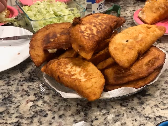 A plate piled with several golden-brown fried pastries on a granite countertop, with a container of shredded lettuce and a bottle of sour cream in the background.