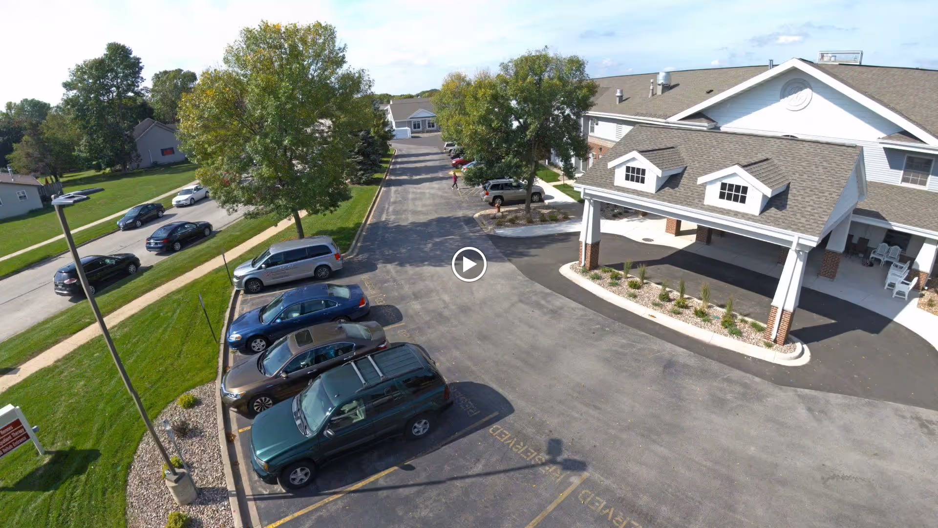 Aerial view of the facility's front entrance and parking lot with a covered porte-cochere, parked cars, lawns and trees.