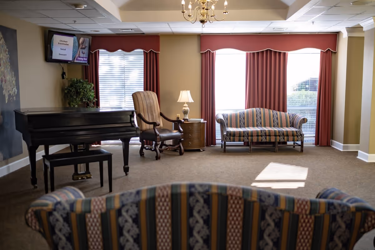 A cozy living room area in a retirement facility featuring a black grand piano with a matching bench, a striped armchair, a wooden side table with a lamp and books, and a patterned sofa. The room has large windows with red curtains and valances, allowing natural light to brighten the space. A chandelier hangs from the ceiling, and a TV mounted on the wall displays a menu with items like Chicken Enchiladas, Salad, and Dessert.
