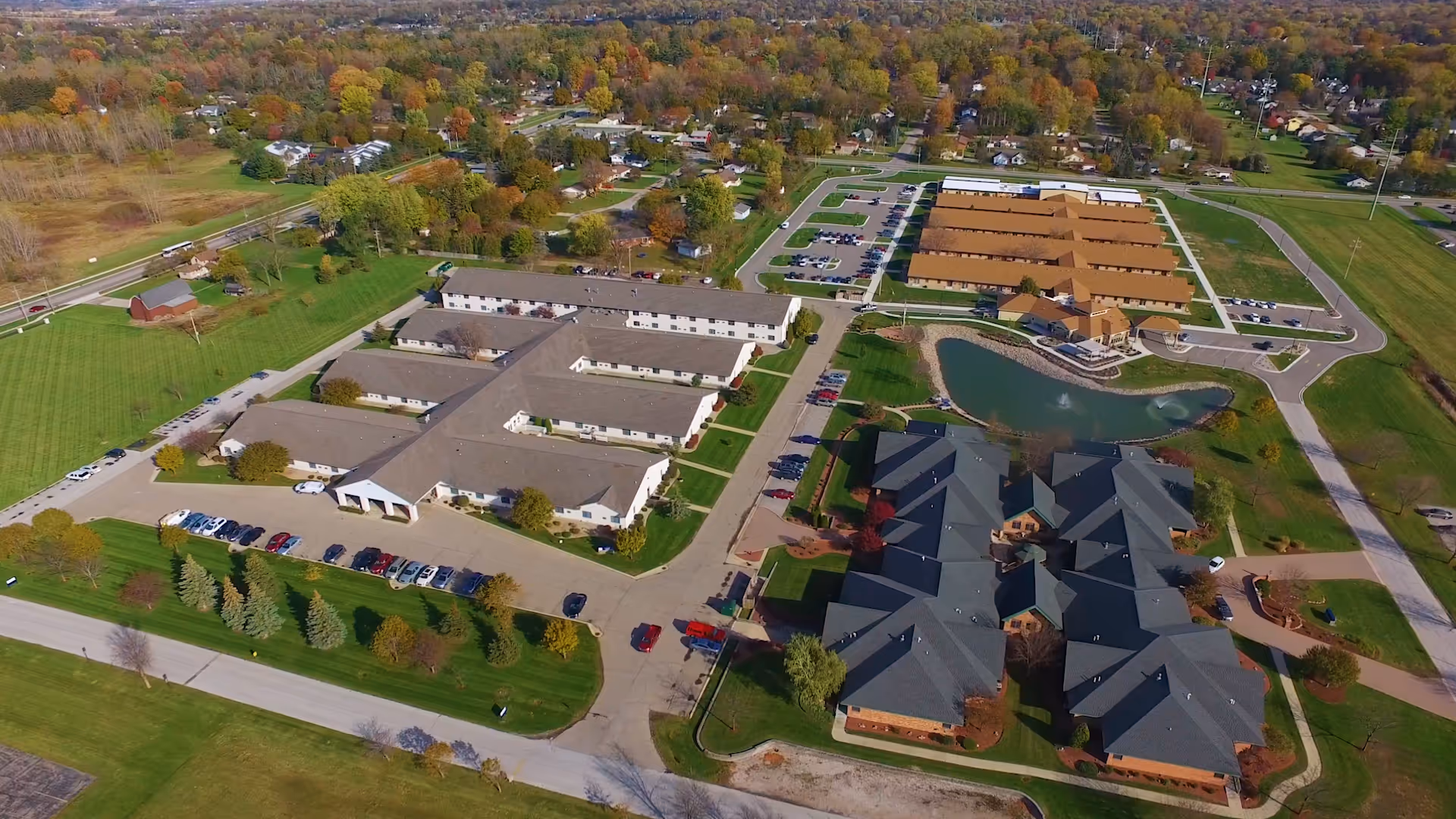 Aerial view of Wellspring Lutheran Services Senior Living Campus showing multiple buildings with parking lots, green lawns, trees, and a pond in the center. The campus is surrounded by residential neighborhoods and open fields.
