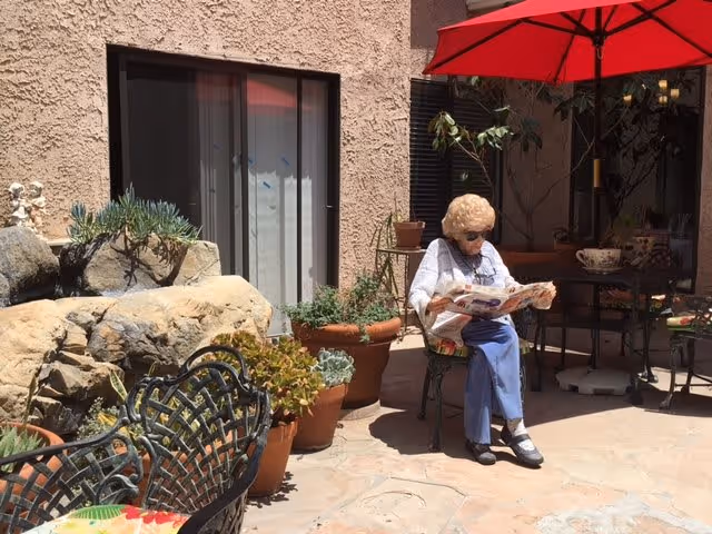 An elderly woman wearing sunglasses sits on a chair under a red patio umbrella, reading a newspaper in a sunny outdoor courtyard with potted plants and rock decorations.