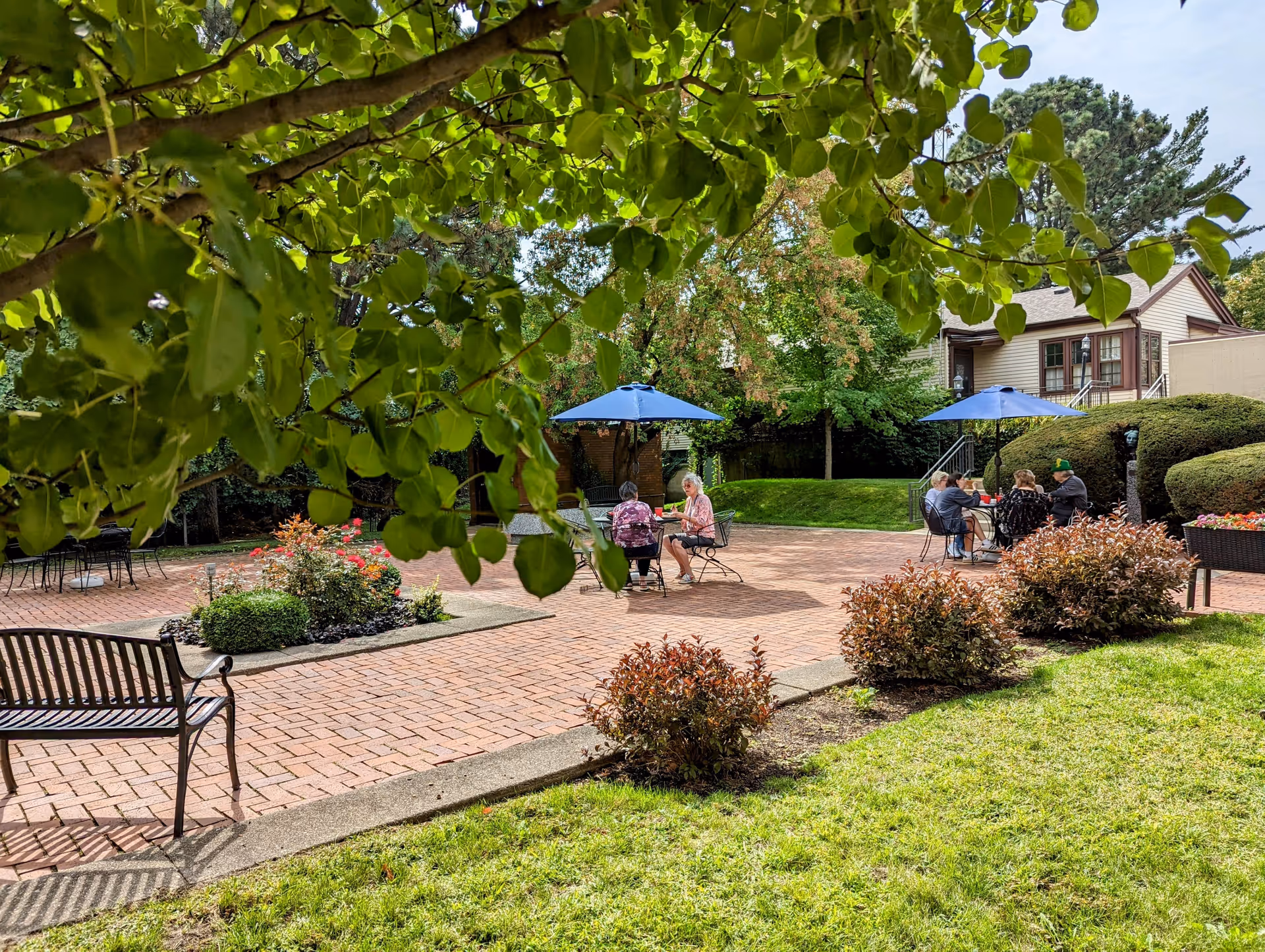 A view of an outdoor patio at Plum Landing, featuring several people sitting at tables under blue umbrellas, surrounded by greenery and flower beds.