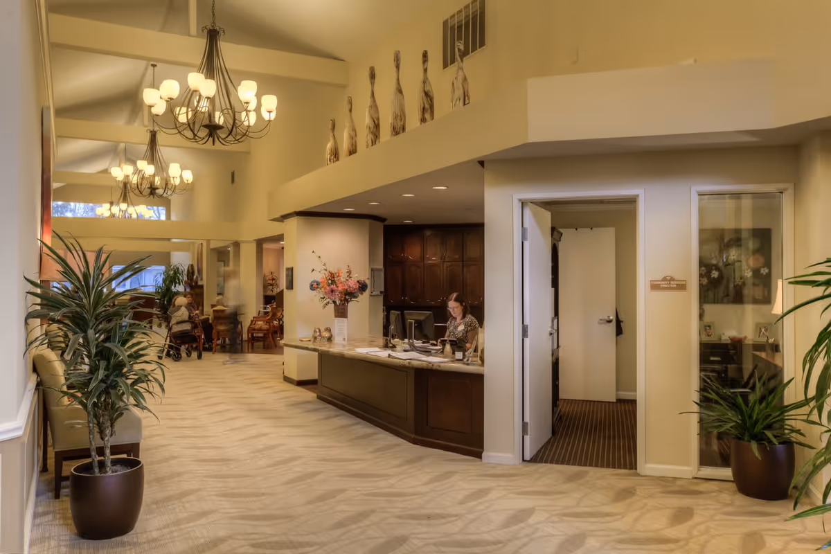 Interior view of a senior living facility reception area with a front desk where a woman is working on a computer. The space features high ceilings with chandeliers, decorative plants, and seating areas along the hallway. There are sculptures displayed on a ledge above the reception desk and a partially open door leading to another room.