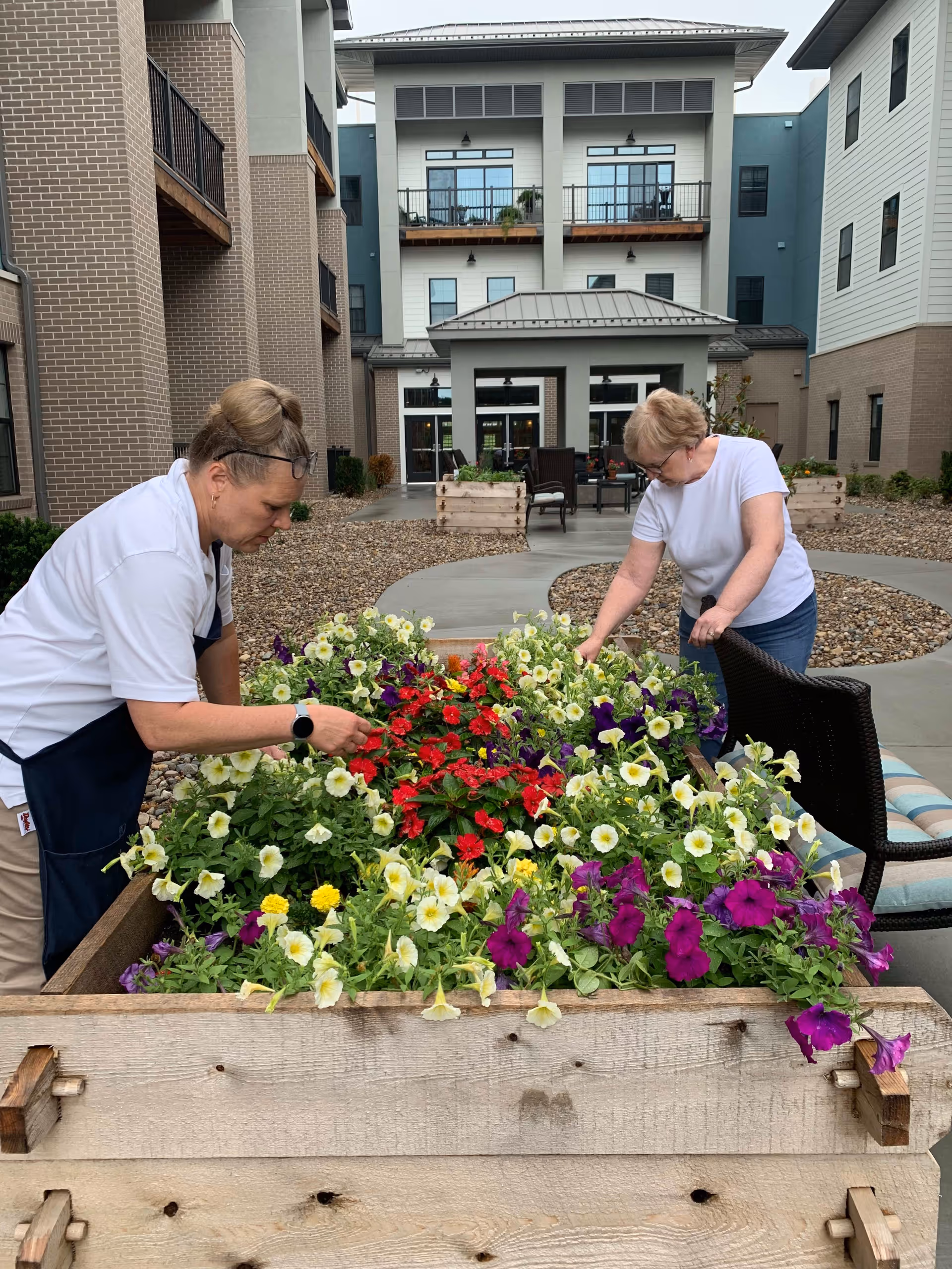 Two women tending to a raised garden bed filled with colorful flowers in an outdoor courtyard area of a senior living facility. The courtyard is surrounded by multi-story buildings with balconies and large windows. There are chairs and planters in the background.