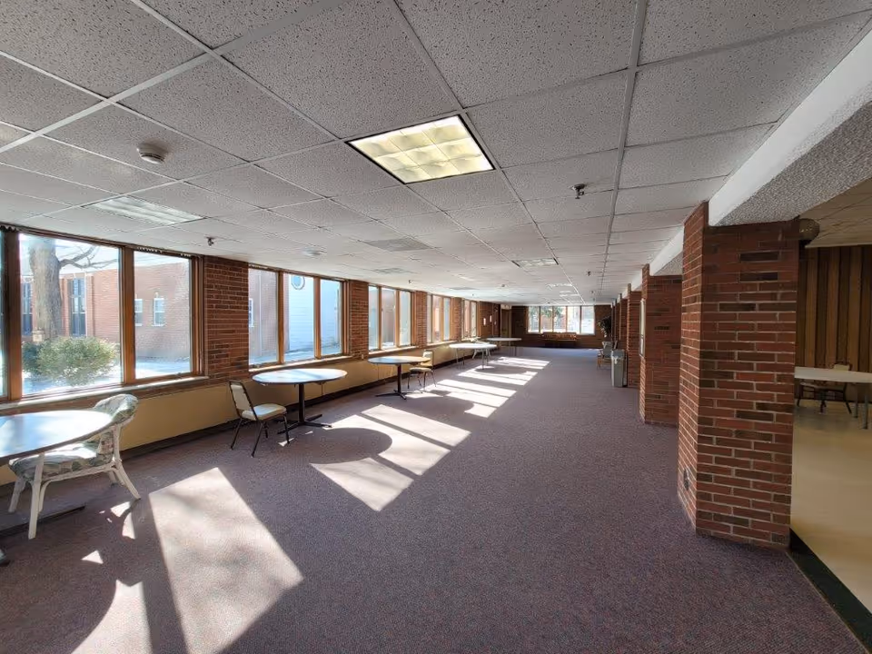 A long, narrow room with large windows along one side letting in natural light. The room has a carpeted floor, brick columns, and several round tables with chairs spaced along the windows. The ceiling has white tiles with fluorescent lighting. Outside the windows, some trees and a brick building are visible.