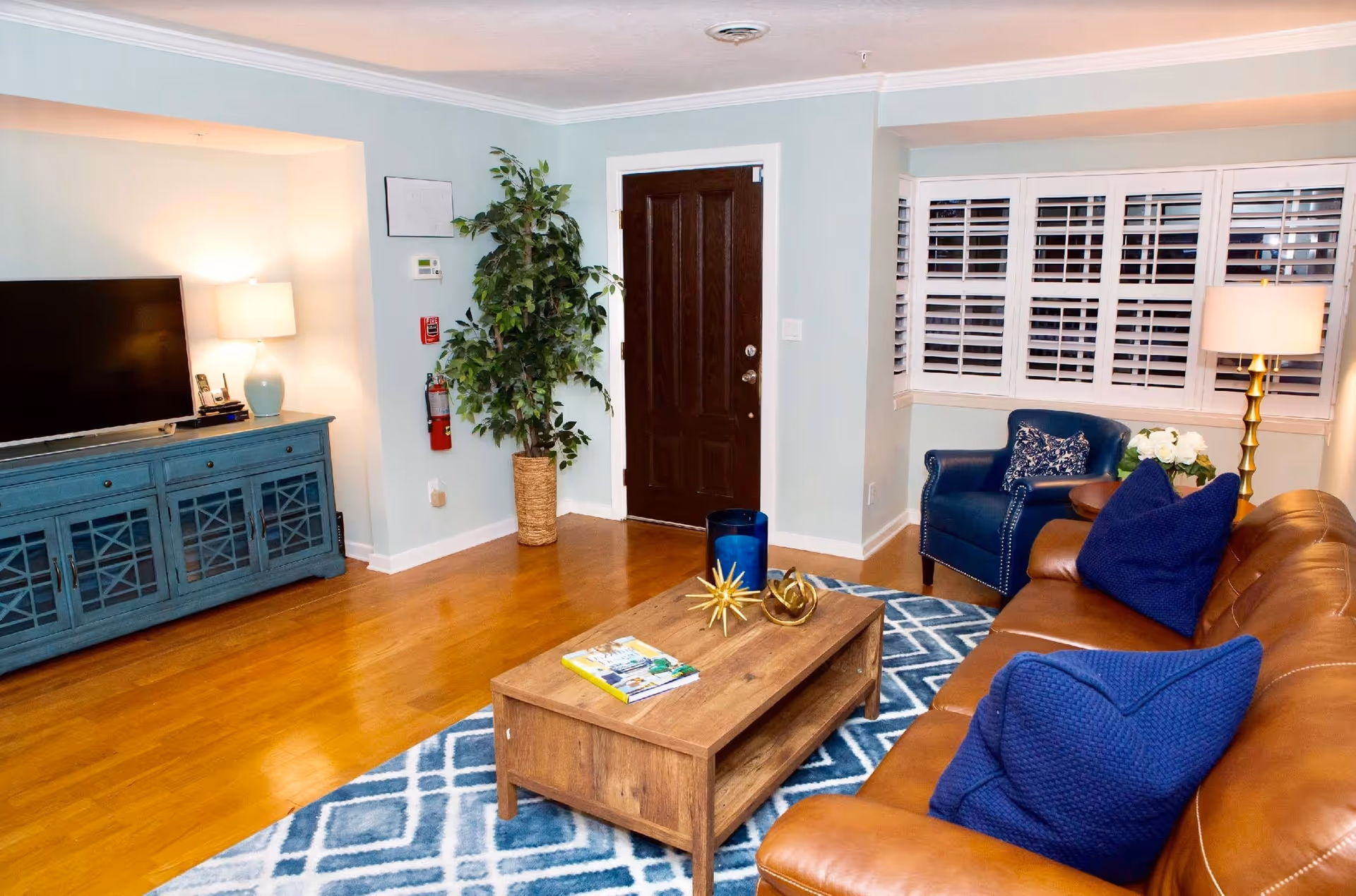 Living room with a brown leather sofa, blue accent chair, wooden coffee table, TV on a blue console, and a dark wood entry door.