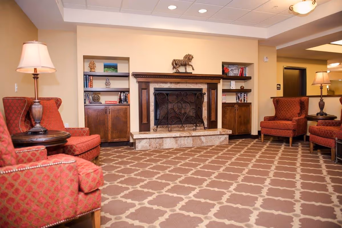 A cozy senior living facility lounge area with patterned carpet, red upholstered armchairs, wooden side tables with lamps, built-in wooden shelves with decorative items, and a fireplace with a decorative horse statue on the mantel.
