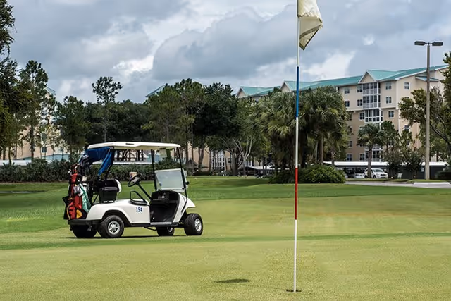 Golf cart parked on a green golf course near a flagstick with a multi-story building and trees in the background under a cloudy sky.