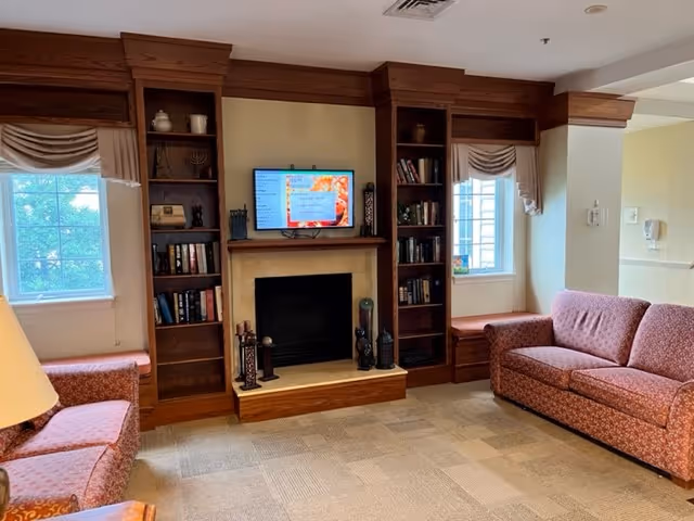 A cozy living room area with two patterned sofas facing a fireplace with a mounted flat-screen TV above it. The fireplace is flanked by built-in wooden bookshelves filled with books and decorative items. Two windows with beige curtains allow natural light into the room. The floor is carpeted with a neutral pattern.