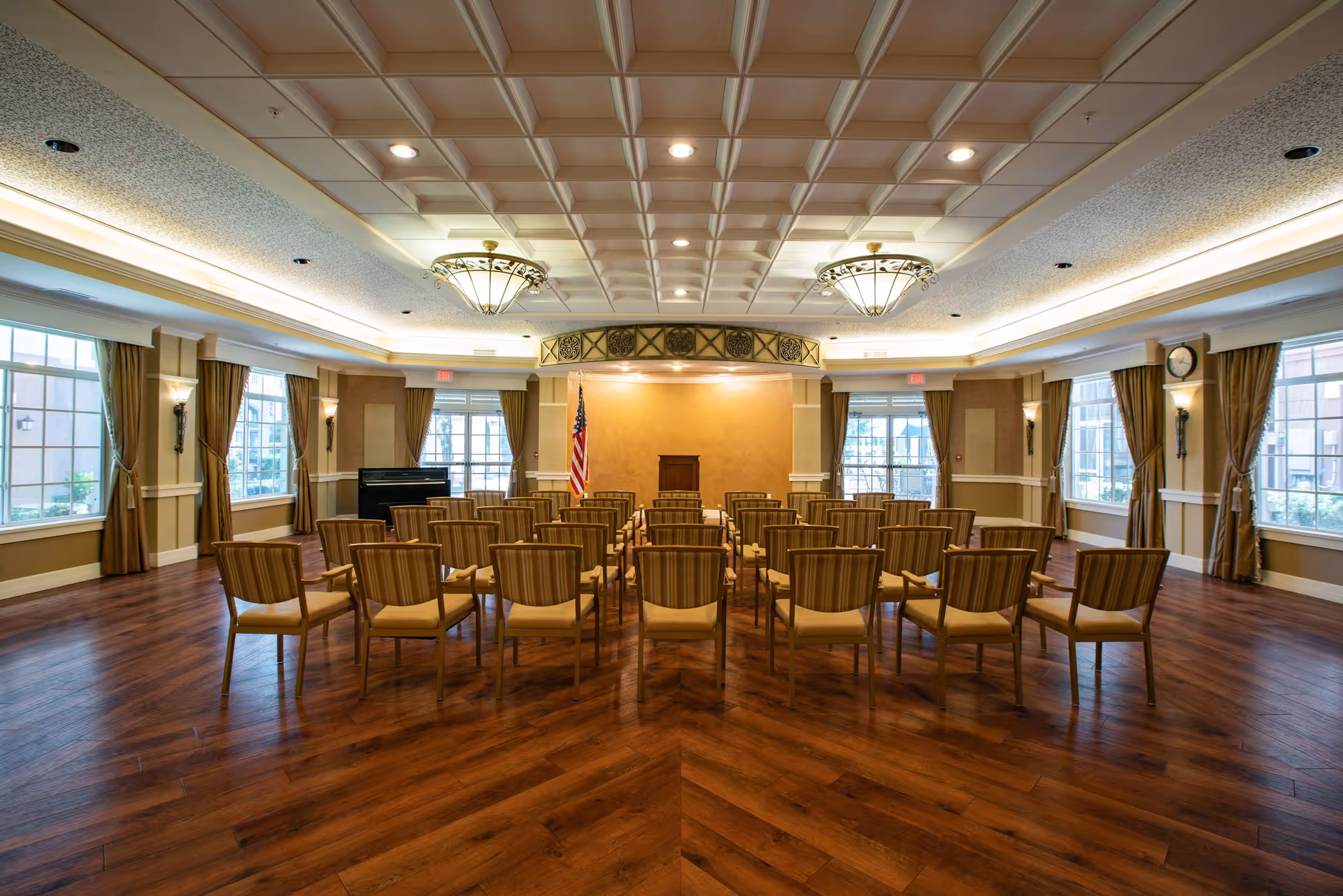 A spacious, well-lit meeting or event room with rows of wooden chairs arranged facing a small podium and an American flag. The room features large windows with brown curtains, a wooden floor, decorative ceiling lights, and a piano in the corner.