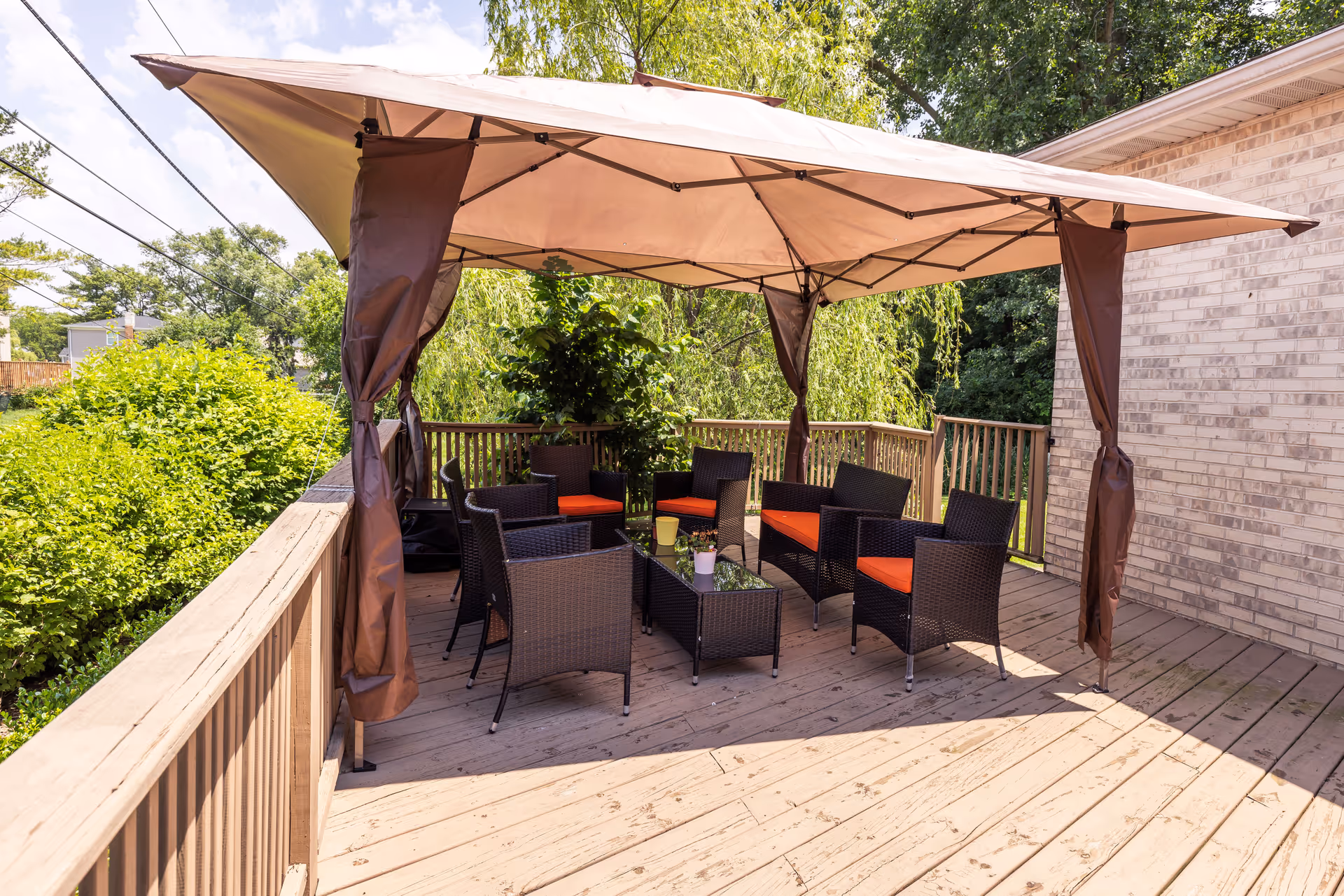 Outdoor wooden deck area with a large beige canopy providing shade over a set of dark wicker chairs and sofas with orange cushions arranged around a glass coffee table. The deck is surrounded by wooden railings and lush green trees and bushes are visible in the background.
