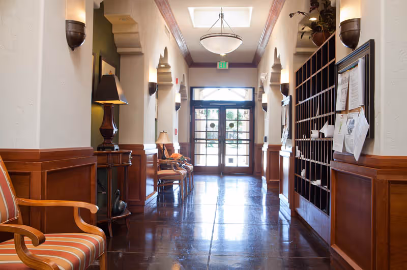 A well-lit hallway in a senior living facility with polished floors, wooden paneling on the walls, wall sconces, and a skylight. There are chairs and a small table with a lamp on the left side, and a mail or message center with papers pinned on a bulletin board on the right side. Glass double doors are visible at the end of the hallway.