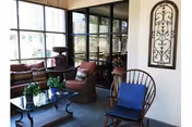 A cozy living room area with large windows letting in natural light. The room features a cushioned sofa with patterned pillows, a glass coffee table with a potted plant, a wooden chair with a blue cushion, and decorative wall art. There are also additional chairs and a small table with flowers near the windows.
