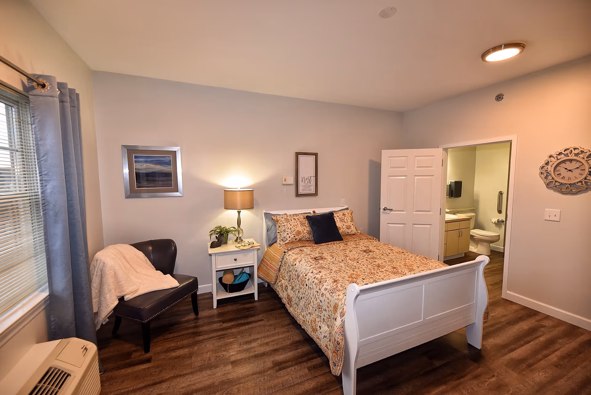 A cozy bedroom in a senior living facility featuring a white wooden bed with floral bedding, a nightstand with a lamp and a small plant, a dark leather chair with a light-colored throw blanket, a window with blue curtains, and an open door leading to a bathroom with a toilet and sink visible.