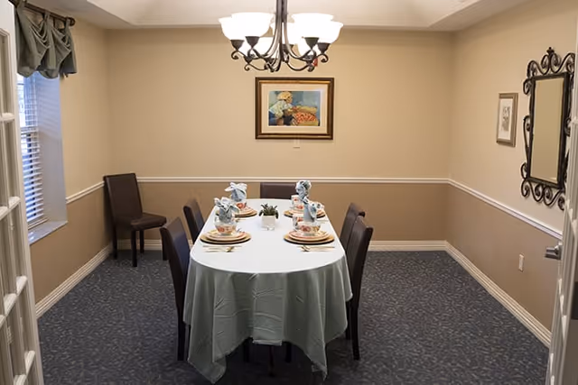 A dining room with a rectangular table covered with a white tablecloth and set for six people with plates, napkins folded in decorative shapes, and glasses. The room has beige walls with a white chair rail, a framed painting on the wall, a decorative mirror, and a chandelier overhead. There are six dark brown chairs around the table and a window with a green valance on the left side.