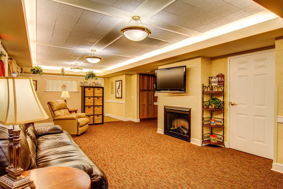 A cozy living room area with a brown leather sofa and a beige recliner chair. There is a wooden side table with a lamp next to the sofa. The room features a wall-mounted flat screen TV above a fireplace, a shelving unit with plants and decorative items, and a cabinet with woven baskets. The ceiling has recessed lighting and decorative light fixtures, and the floor is carpeted in a warm pattern.