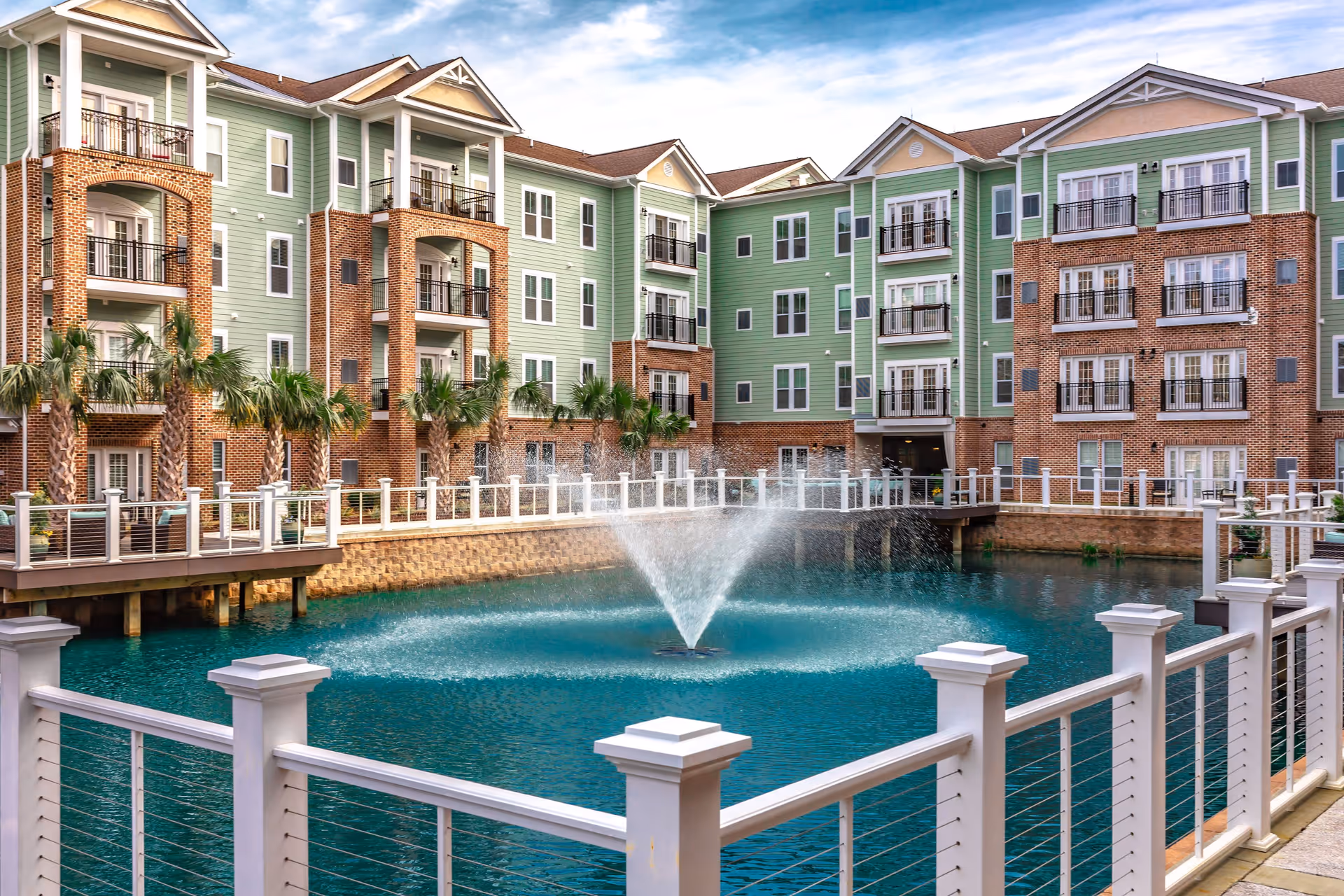 View of a multi-story residential building with green siding and brick accents surrounding a large pond with a central water fountain, bordered by a white railing and palm trees.