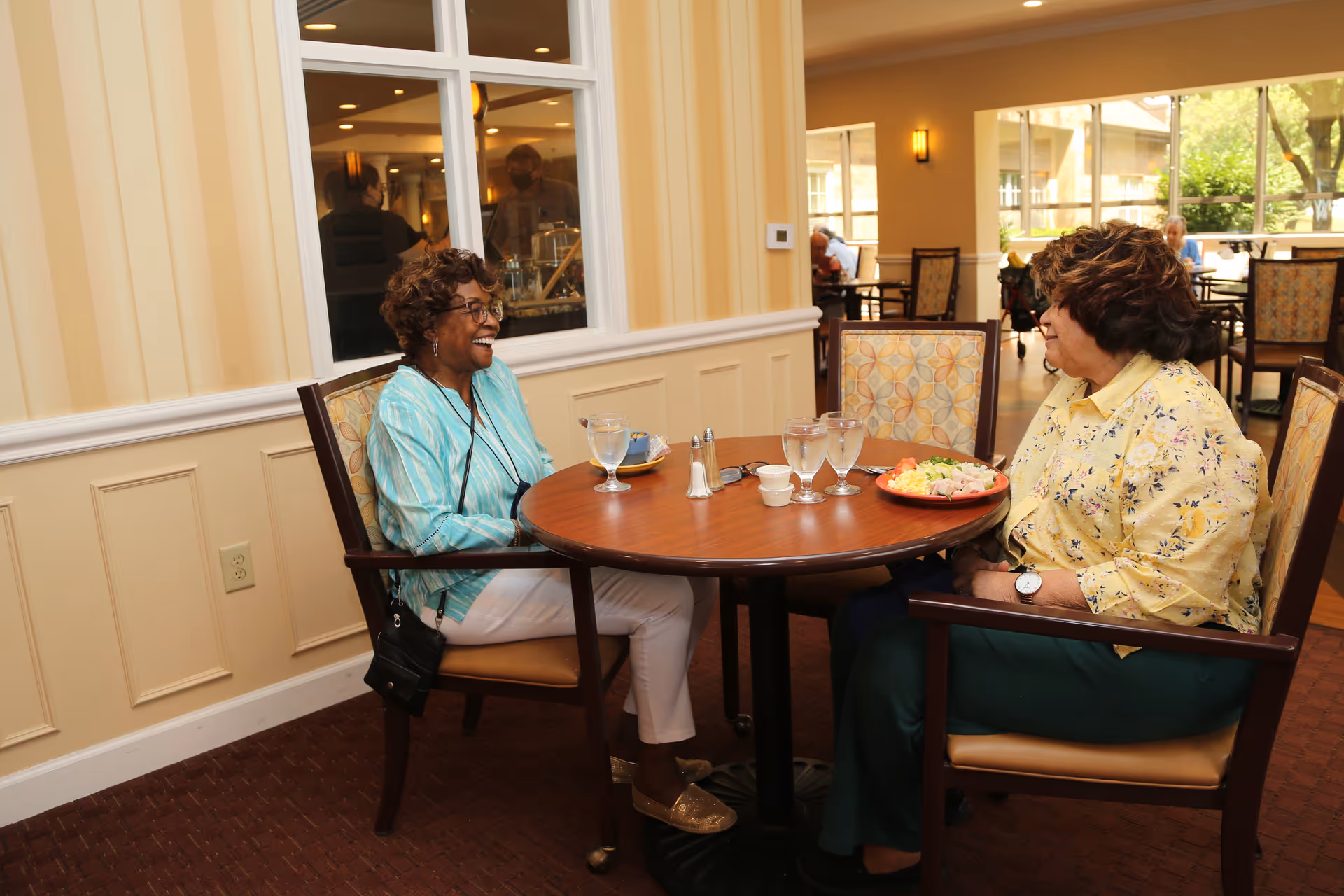 Two elderly women sitting at a round wooden table in a dining area, smiling and engaged in conversation. One woman is wearing a light blue striped shirt and white pants, while the other is wearing a yellow floral blouse and green pants. The table has glasses of water, a plate of food, and salt and pepper shakers. The background shows more tables and chairs with other people dining, and large windows letting in natural light.