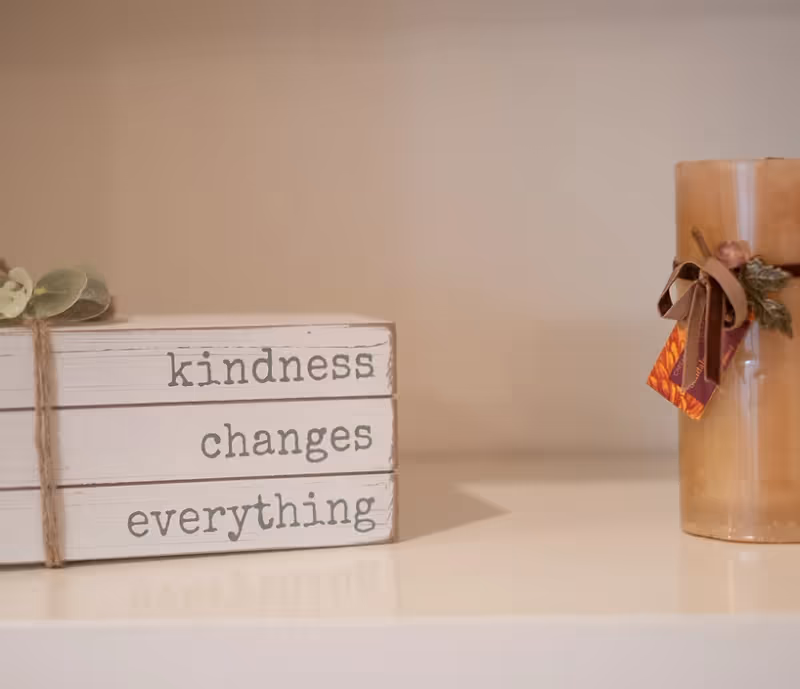 A decorative stack of three white wooden blocks with the words 'kindness changes everything' printed on them, next to a large beige candle adorned with a ribbon and small decorative leaves, placed on a white surface against a plain beige background.