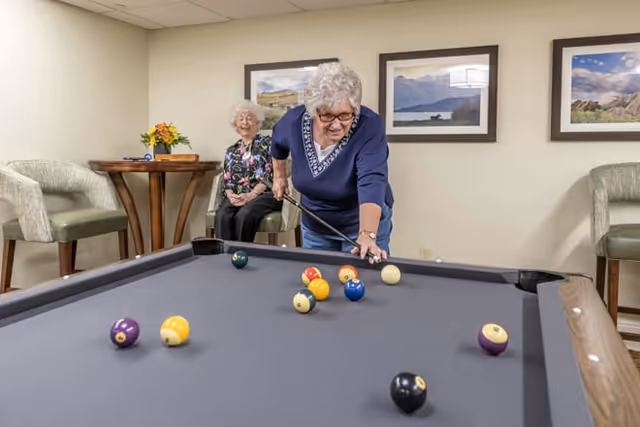 Two elderly women in a room with a pool table. One woman is leaning over the table preparing to take a shot, while the other woman is seated in the background smiling. The room has framed landscape pictures on the wall and comfortable chairs around a small table with a flower arrangement.