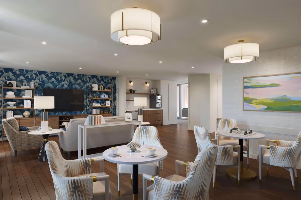 A bright communal lounge with round tables and upholstered chairs, sofas, a wall-mounted TV, bookshelves and decorative lighting.