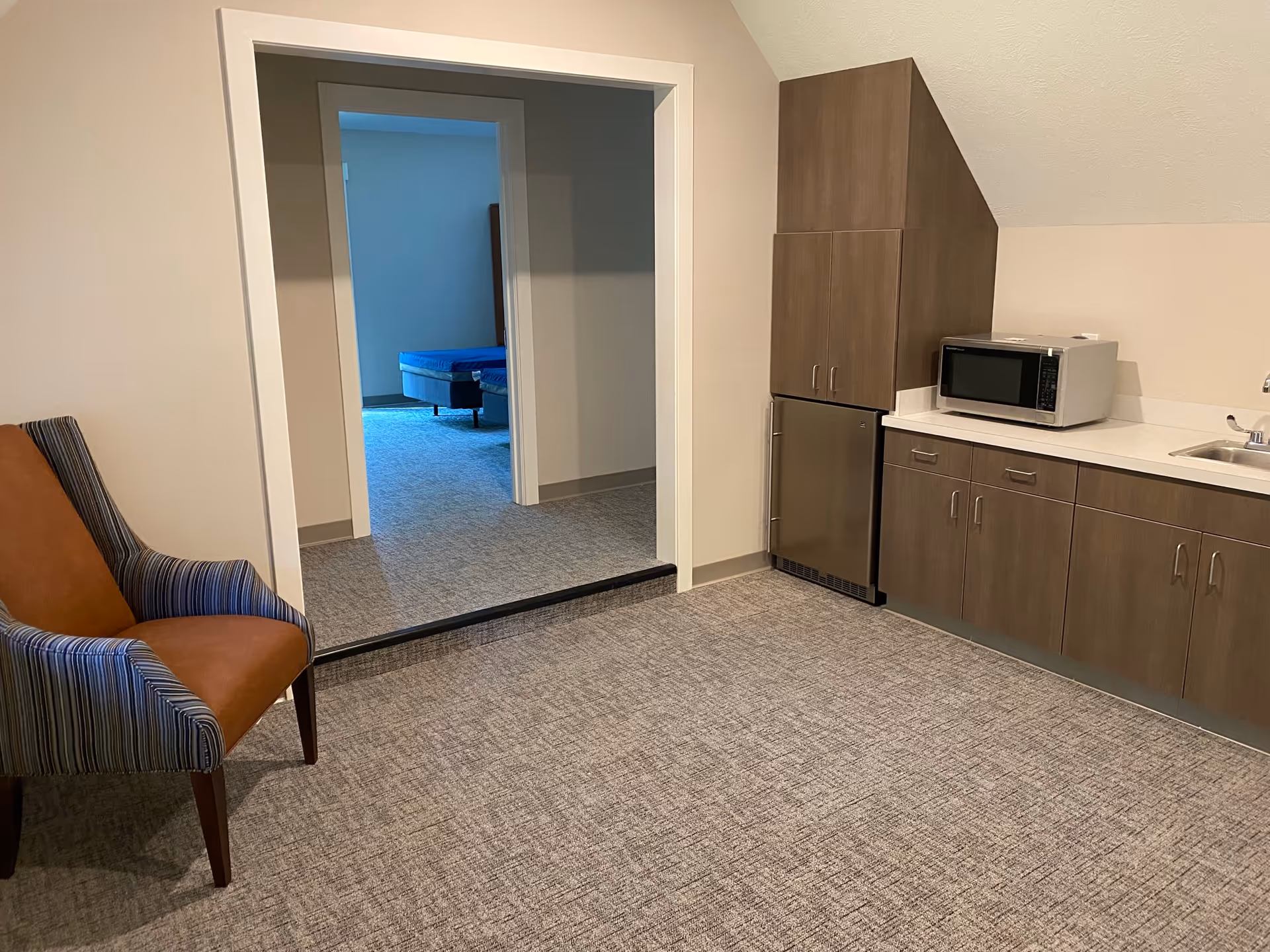 Interior room with a brown and blue striped armchair on the left, a kitchenette on the right with wooden cabinets, a small refrigerator, a microwave, and a sink. There is an open doorway leading to another room with a bed visible in the background.