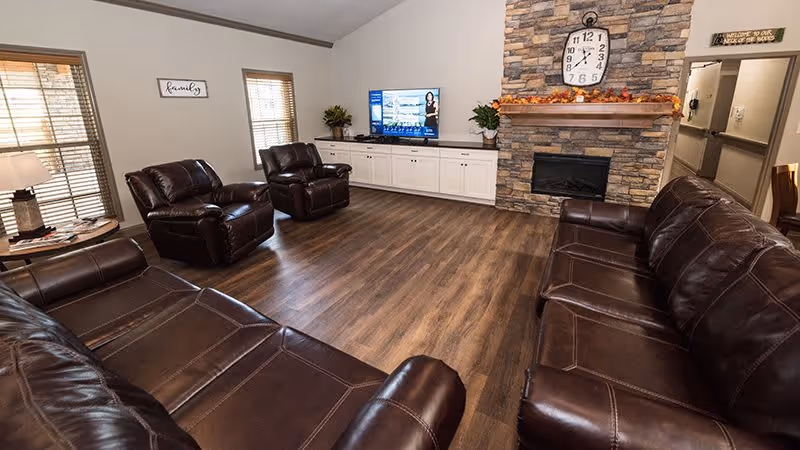 A cozy living room with dark brown leather sofas and armchairs arranged around a wooden floor. A stone fireplace with a large clock above it is decorated with autumn leaves. A flat-screen TV is mounted on a white cabinet against the wall. There are windows with blinds and a lamp on a side table, and a hallway is visible in the background.