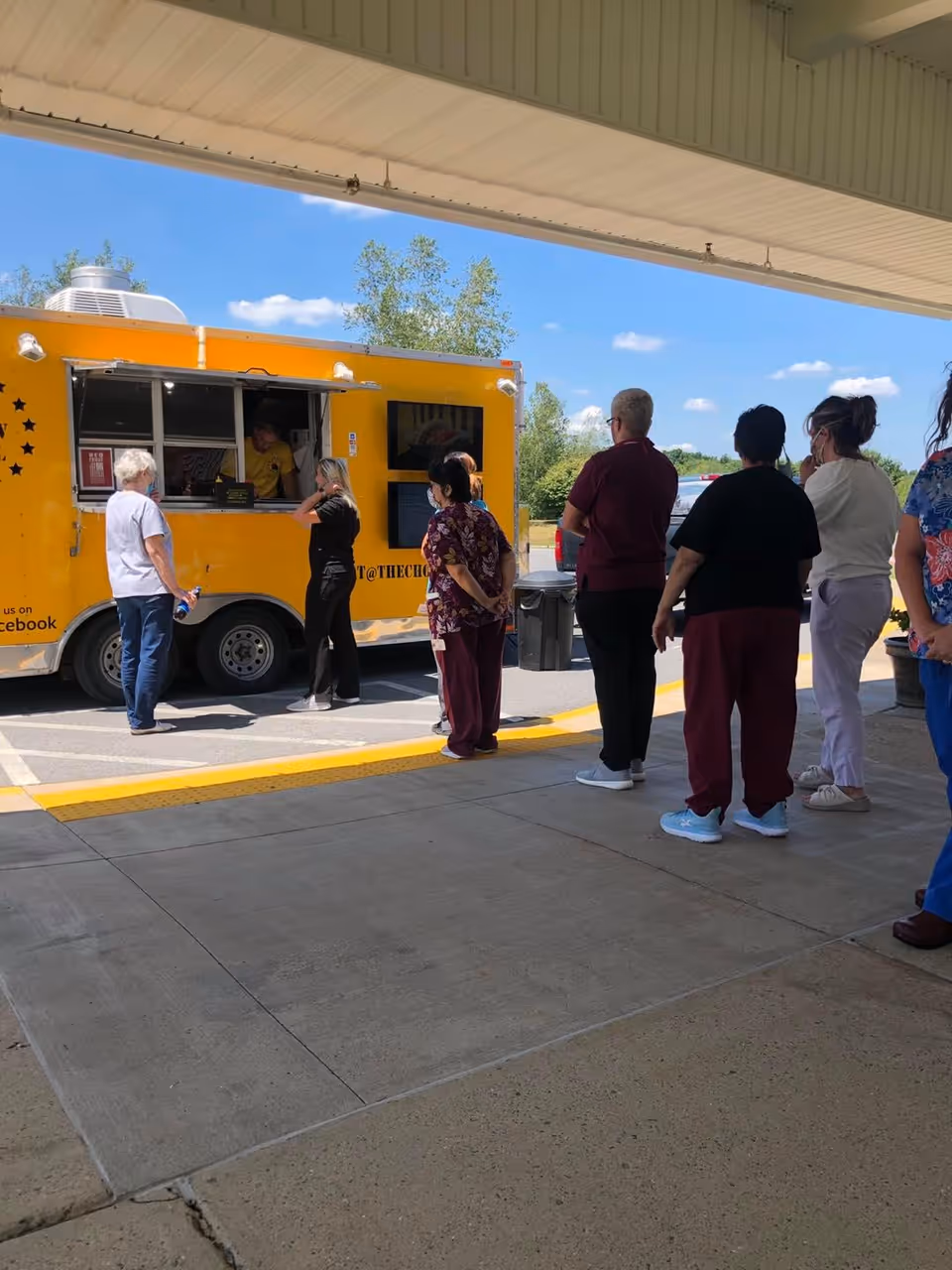 A group of people standing in line outside under a covered area, waiting to order from a bright yellow food truck on a sunny day with a clear blue sky and some trees in the background.